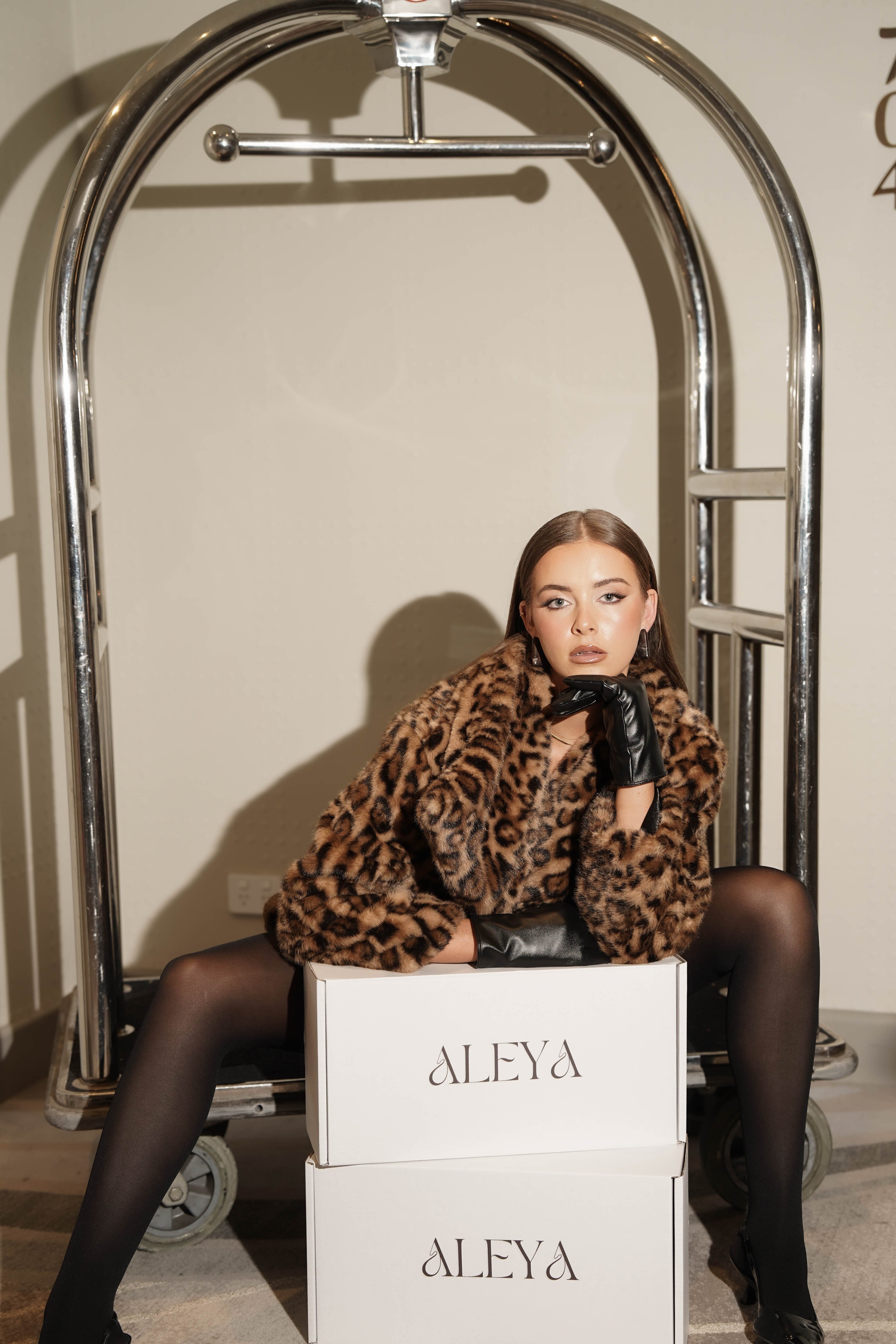 Woman in Aleya Monaco Shopping leopard print coat sitting on 'ALEYA' boxes with a silver luggage cart in the background.